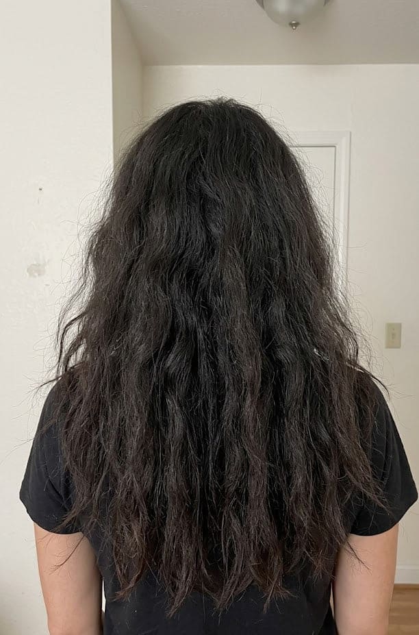 A "Before" photo of a person from behind, showing long, dark, wavy hair that appears frizzy, dry, and lacking definition. The hair has significant volume but looks dull and unmanaged against a plain white wall.