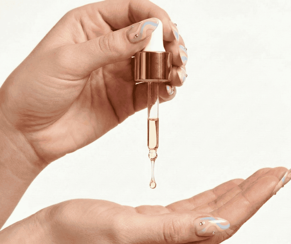 Rose gold dropper bottle with a hand holding it over another hand on a light background