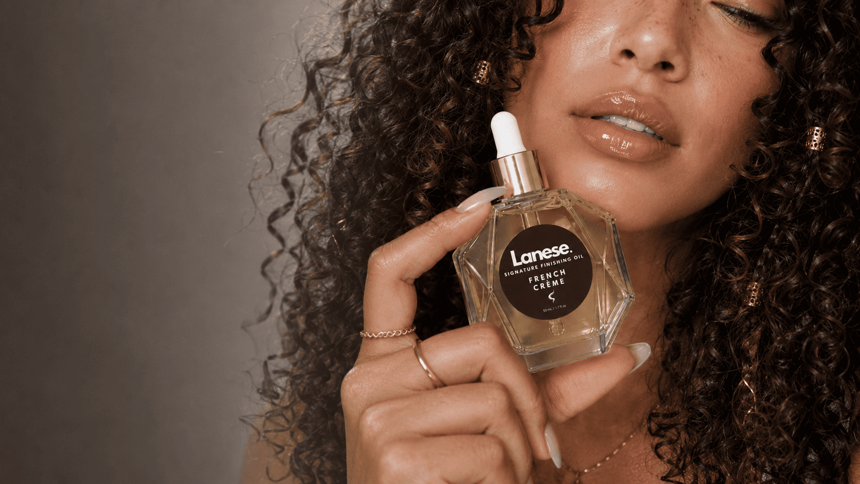 A close-up lifestyle shot of a woman holding a bottle of Lanese. The woman has voluminous curly dark brown hair and is holding the clear glass bottle which contains a golden liquid, featuring a white dropper cap and a black circular label.