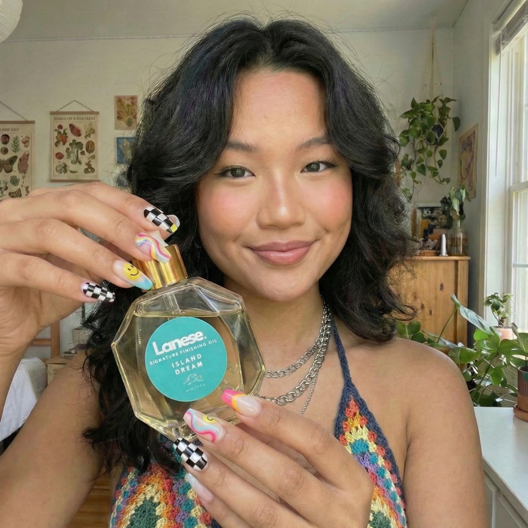 A smiling woman with dark, wavy shoulder-length hair wearing a colorful crochet halter top, holding up a hexagon-shaped glass dropper bottle of Lanese hair oil. Her nails feature fun, colorful checkerboard and smiley face designs. The teal label on the bottle reads: "Lanese. SIGNATURE FINISHING OIL. ISLAND DREAM. 50 mL / 1.7 fl oz".