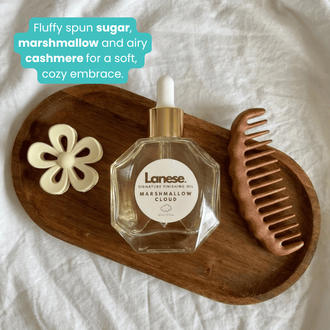 A top-down lifestyle shot of a Lanese Marshmallow Cloud bottle sitting on an oval wooden tray alongside a white flower hair claw and a brown wide-tooth comb. A teal text box describes the scent: "Fluffy spun sugar, marshmallow and airy cashmere for a soft, cozy embrace."