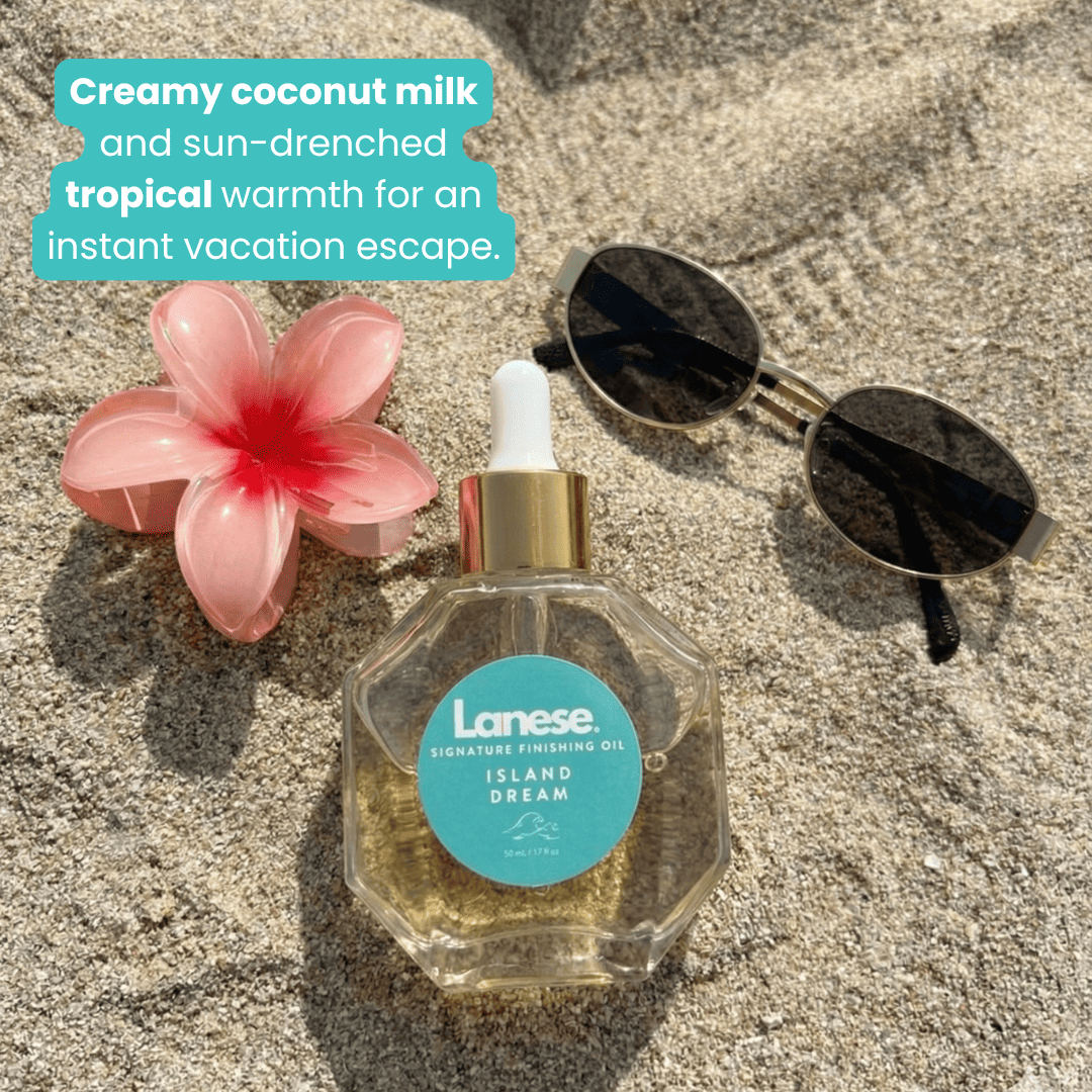 A lifestyle shot of the Lanese Island Dream oil bottle sitting in the sand at a beach next to a pink flower hair clip and black sunglasses. A teal text bubble at the top left says: "Creamy coconut milk and sun-drenched tropical warmth for an instant vacation escape."