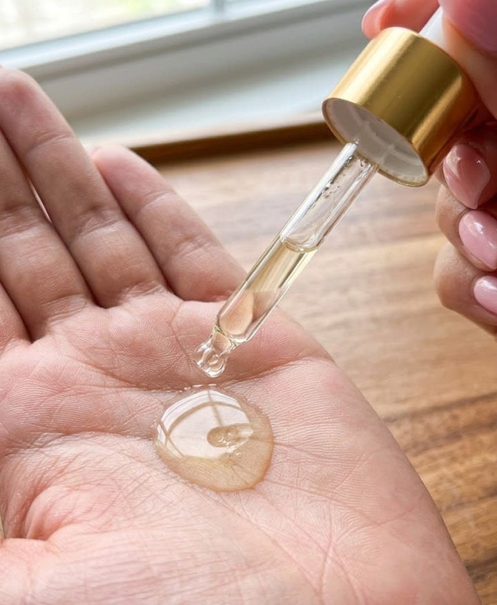 Gold dropper bottle with clear liquid being applied to a hand on a wooden surface.